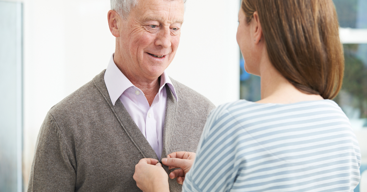 Family Caregiver Buttoning The Sweater Of An Elderly Gentleman.