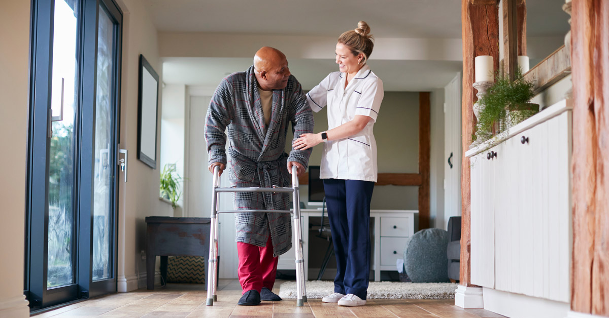 Nurse Assisting An African Man With His Walker In His Home.